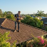 Professional roofer in safety gear inspecting ridge caps and tiles on a suburban Brisbane home roof under clear spring skies, emphasizing proactive maintenance.