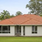 Single-story house with a red tiled roof, light exterior walls, and a green lawn, surrounded by palm trees and tall trees.