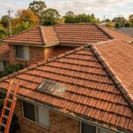 Tiled house roof with tools and ladder set up for repairs in a suburban neighborhood.
