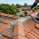 A roofer applying fresh grey mortar to the ridge of a terracotta tiled roof in a suburban area.